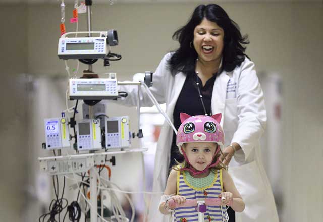 Smiling girl riding bike in hallway, hooked up to healthcare machine while smiling medical professional pushes machine to keep up