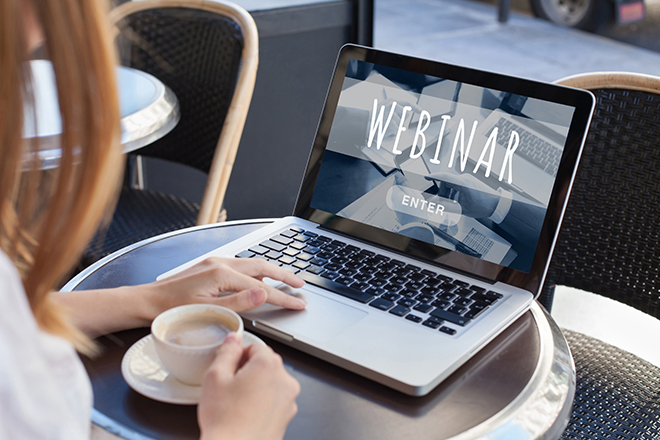 A woman sits at a coffeshop, watching a webinar on her laptop.