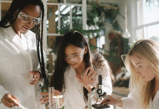 stock photo of a black woman with braids, an asian woman and a blonde woman wearing clear safety goggles and white lab coats huddled around various lab equipment 